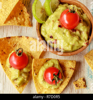 Les croustilles de maïs et de guacamole nachos dans un bol en bois décoré de tomates cerises et des tranches de citron vert sur un tableau blanc en bois. Banque D'Images