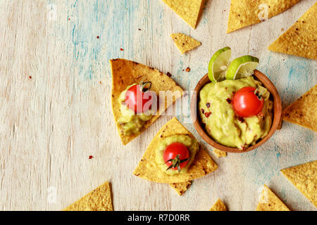 Les croustilles de maïs et de guacamole nachos dans un bol en bois décoré de tomates cerises et des tranches de citron vert sur un tableau blanc en bois. Eith espace. Banque D'Images