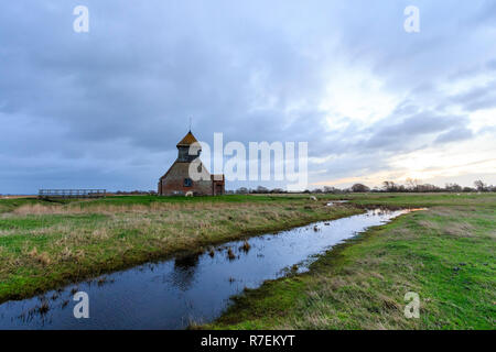 Lever du soleil dans un ciel nuageux sur l'Eglise sur le Marais, église St Thomas Becket, à Fairfield, Romney Marsh dans le Kent. L'église du xviiie siècle, construit autour d'une ancienne église du xie siècle, se trouve seul sur le marais, entourée de fossés de drainage et les moutons. Banque D'Images