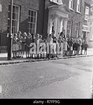 1965, un groupe d'enfants de l'école primaire debout sur le côté de la route en attendant de voir une parade aller passe-haut, St, Aylesbury, Angleterre, Royaume-Uni. Banque D'Images