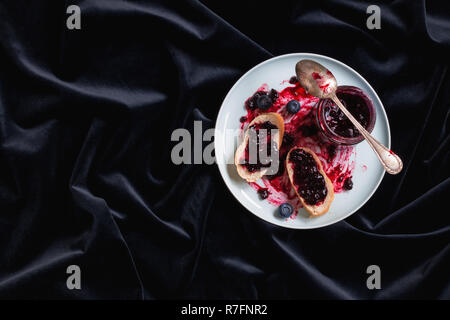 Pot de confiture de bleuets, à côté de pain sur une plaque. Le tout sur un fond de velours noir. Place pour le texte. Banque D'Images