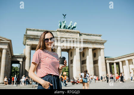 Une fille est titulaire d'un gobelet jetable avec du café ou une autre boisson lors de la marche ou de visites à Berlin, Allemagne. L'avant est la porte de Brandebourg et de personnes non reconnu floue. Banque D'Images