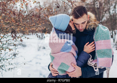 Beau couple aimant la marche et s'étreindre dans la forêt d'hiver. Personnes couvertes de réchauffement d'une couverture. Woman whispering quelque chose à son petit ami Banque D'Images
