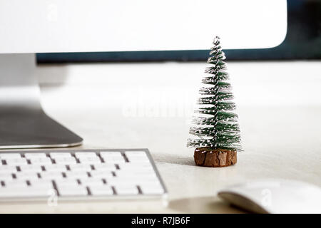 Un petit sapin se dresse sur un bureau blanc, près de l'écran d'un ordinateur, clavier et souris. Concept, créer une ambiance de Noël sur le lieu de travail. Banque D'Images