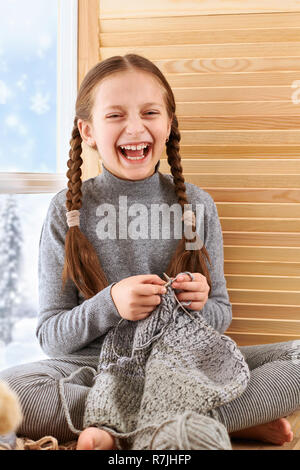 Fille de l'enfant est assis sur un rebord de fenêtre et tricoter son écharpe de laine grise. Belle vue sur l'extérieur de la fenêtre - journée ensoleillée en hiver et de la neige. Banque D'Images