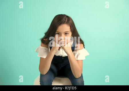 Girl prop menton sur les mains sur fond bleu. Enfant avec de longs cheveux de brunette en jeans dans l'ensemble. Beauté, mode, look, concept, copy space Banque D'Images