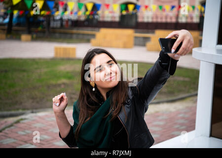 Dreamy brunette girl exprimant des émotions heureux tout en faisant l'extérieur selfies. Photo de jeune femme élégante de prendre photo d'elle-même sur l'arrière-plan flou. Banque D'Images