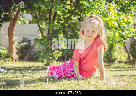 Adorable petite fille blonde avec la cerise dans les oreilles assis dans l'herbe verte Banque D'Images