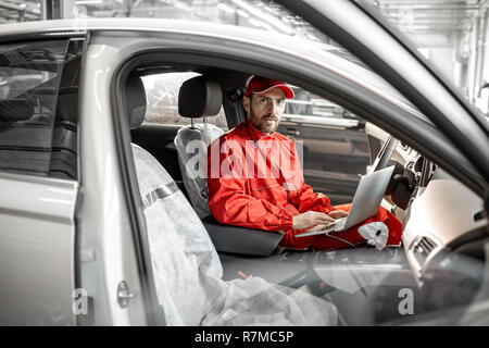 Mécanicien Auto en uniforme rouge diagnostic voiture avec ordinateur assis sur le siège conducteur de la voiture publique Banque D'Images