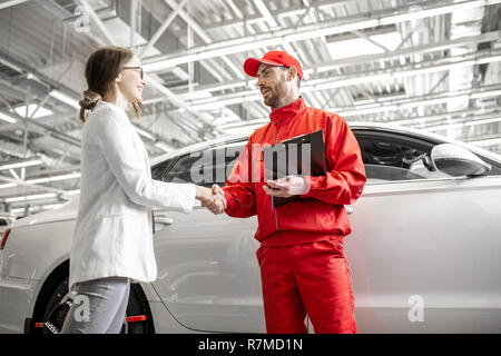 Young woman shaking hands with client mécanicien auto en uniforme rouge ayant un accord à l'car service Banque D'Images