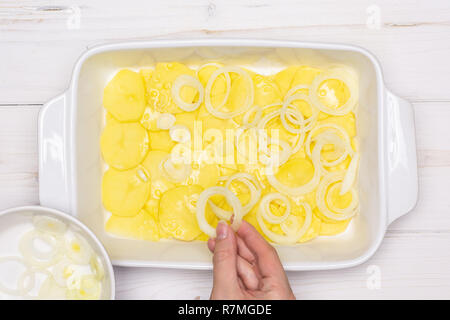 Femme mains mettent des couches de l'oignon cru tranches. étape par étape de recette pommes de terre au four avec l'oignon dans une cocotte en céramique blanche sur flatlay bois blanc Banque D'Images