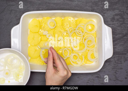 Femme mains mettent des couches de l'oignon cru tranches. étape par étape de recette pommes de terre au four avec l'oignon dans une cocotte en céramique blanche en pierre grise sur flatlay Banque D'Images