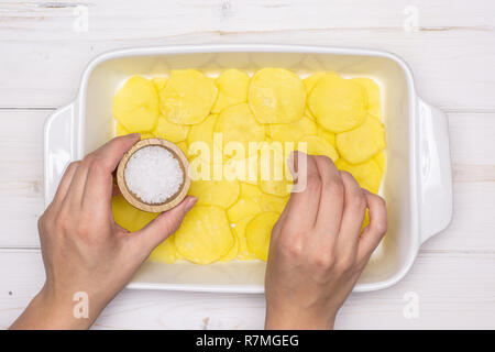 Femme mains sont le salage couches. Recette étape par étape, les pommes de terre cuites au four avec l'oignon dans une cocotte en céramique blanche sur flatlay bois blanc Banque D'Images