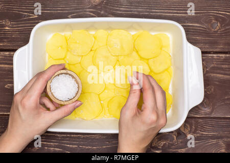 Femme mains sont le salage couches. Recette étape par étape, les pommes de terre cuites au four avec l'oignon dans une cocotte en céramique blanche sur flatlay brun bois Banque D'Images