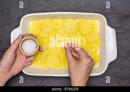 Femme mains sont le salage couches. Recette étape par étape, les pommes de terre cuites au four avec l'oignon dans une cocotte en céramique blanche en pierre grise sur flatlay Banque D'Images