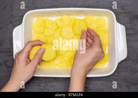 Femme mains mettent des couches de rondelles de pommes de terre recette brute. étape par étape, les pommes de terre cuites au four avec l'oignon dans une cocotte en céramique blanche en pierre grise sur flatlay Banque D'Images