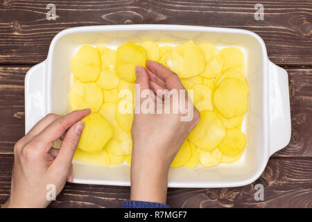 Femme mains mettent des couches de rondelles de pommes de terre recette brute. étape par étape, les pommes de terre cuites au four avec l'oignon dans une cocotte en céramique blanche sur flatlay brun bois Banque D'Images