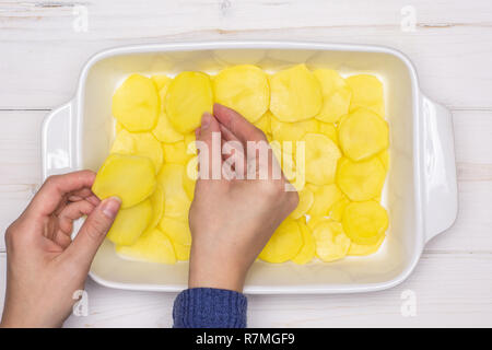 Femme mains mettent des couches de rondelles de pommes de terre recette brute. étape par étape, les pommes de terre cuites au four avec l'oignon dans une cocotte en céramique blanche sur flatlay bois blanc Banque D'Images