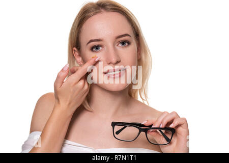 Jeune blond woman holding contact lens sur le doigt devant son visage et tenant dans son autre main un lunettes noires sur fond blanc. Banque D'Images