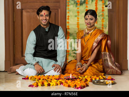 Heureux couple maharashtrian en costume traditionnel assis près de l'entrée avec des décorations de fleurs et de la plaque de pooja diya sur le sol. Banque D'Images