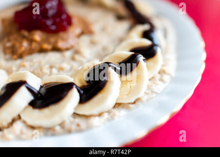 Gros plan macro de grain d'avoine porridge d'avoine pudding bol avec des morceaux de tranches de banane, sauce chocolat arrosé, le beurre d'arachide et confiture, table rose Banque D'Images