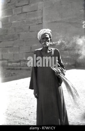 Années 1950, historique, un vieil homme égyptien portant une robe traditionnelle et turban fumer une cigarette à l'extérieur, de l'Égypte. Banque D'Images