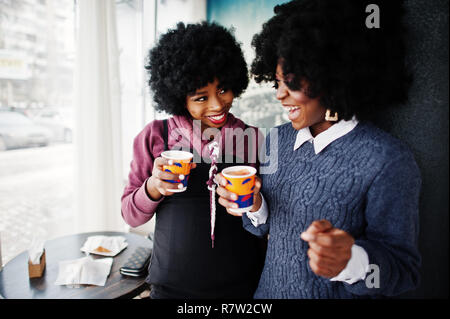 Deux cheveux bouclés african american woman l'usure sur les chandails avec des tasses de thé posée au café à l'intérieur. Banque D'Images