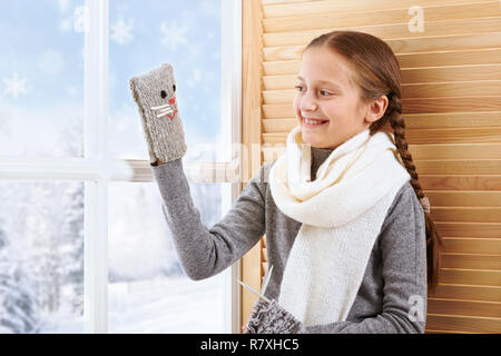 Fille de l'enfant est assis sur un rebord de fenêtre et jouer avec des moufle. Belle vue sur l'extérieur de la fenêtre - journée ensoleillée en hiver et de la neige. Banque D'Images