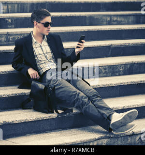 Beau jeune homme d'affaires à l'aide de smart phone on modèle masculin élégant vêtu de noir jacket and sunglasses Banque D'Images