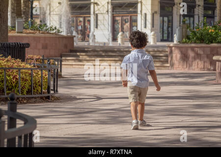 Un petit garçon s'éloigne de l'autre côté de la cour sur une journée ensoleillée. Vêtu de chaussures de tennis, short kaki et une chemise rayée bleu. Banque D'Images