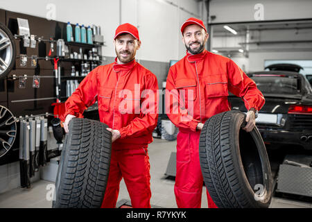 Portrait d'un service de location de deux travailleurs en uniforme rouge se tenant ensemble avec de nouveaux pneus au magasin Banque D'Images