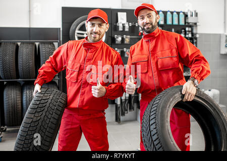 Portrait d'un service de location de deux travailleurs en uniforme rouge se tenant ensemble avec de nouveaux pneus au magasin Banque D'Images