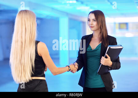 Deux filles dans le bureau avec les dossiers avec des documents en main. Les jeunes femmes se serrent la main. Hall vide bleu sur l'arrière-plan Banque D'Images