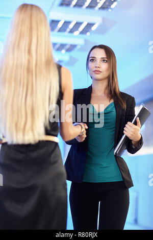 Deux filles dans le bureau avec les dossiers avec des documents en main. Les jeunes femmes se serrent la main. Hall vide bleu sur l'arrière-plan Banque D'Images