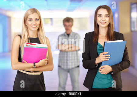 Deux filles dans le bureau avec les dossiers avec des documents en main. Les jeunes femmes sont bavardant et riant. Leur patron en colère regarde de derrière. Banque D'Images