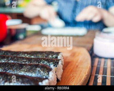 Faire de rouleaux de sushi : Étanchéité des rouleaux sur la planche de bois avant de trancher. Femme floue mains mettre du riz sur une feuille de nori et ustensiles en arrière-plan. Banque D'Images