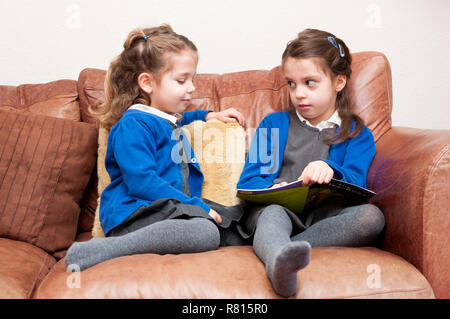 Les jeunes sœurs en uniforme de l'école primaire la lecture d'un livre ensemble, assis sur un canapé, Royaume-Uni Banque D'Images