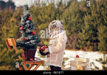 Un enfant décorer un arbre de Noël à l'extérieur. Le garçon se bloque et bulles rouge mange gingerbreads. Il est habillé d'un manteau en peau de mouton vert, dans l'arrière-plan Banque D'Images