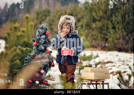 Un enfant décorer un arbre de Noël à l'extérieur. Le garçon se bloque et bulles rouge mange gingerbreads. Il est habillé d'un manteau en peau de mouton vert, dans l'arrière-plan Banque D'Images