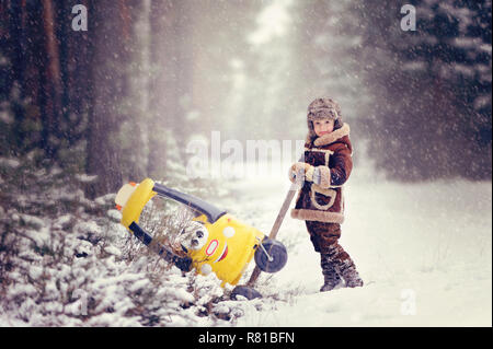 Enfant jouant à l'extérieur au cours de neige. Le garçon pousse de la neige petite voiture jaune, avec deux ours en peluche à l'intérieur, belle hiver neigeux par Banque D'Images