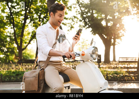 Cheerful young businessman assis sur une moto à l'extérieur, à l'aide de mobile phone Banque D'Images