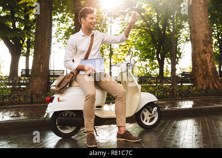 Cheerful young businessman assis sur une moto à l'extérieur, travailler sur ordinateur portable, en agitant la main Banque D'Images