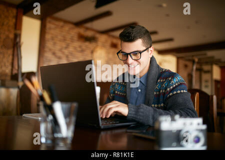 Smiling attractive hipster homme dans les verres et uncategorized working on laptop in cafe. La navigation sur Internet à l'homme d'affaires indépendant café. Retouche photographe photos. Vintage camera sur table. Banque D'Images