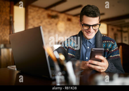 Un jeune homme en tapant un texte sur mobile smartphone moderne. Hipster tenant un téléphone moderne et l'écriture d'un message téléphonique. Smiling young woman in glasses looking at cellphone avec ordinateur portable sur la table. Banque D'Images