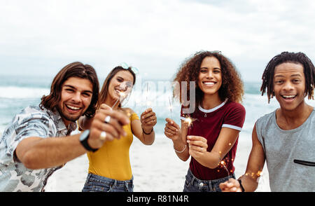 Groupe varié de jeunes célébrant le nouvel an sur la plage. Les jeunes hommes et femmes s'amusant avec les cierges magiques en plein air au bord de la mer. Banque D'Images