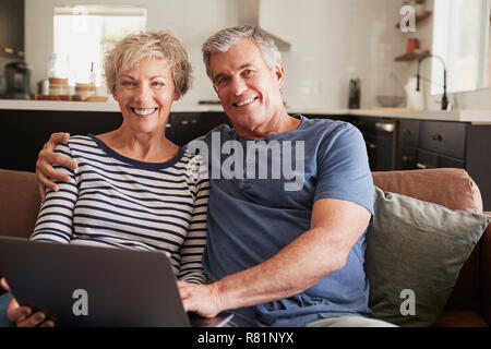 Senior couple sitting on couch, smiling utilisation ordinateur portable à l'appareil photo Banque D'Images