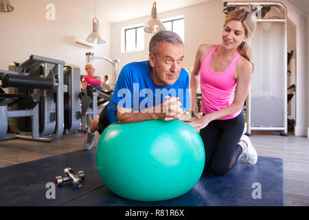 Senior Man Exercising sur Ballon d'être encouragés par l'entraîneur personnel dans la salle de sport Banque D'Images