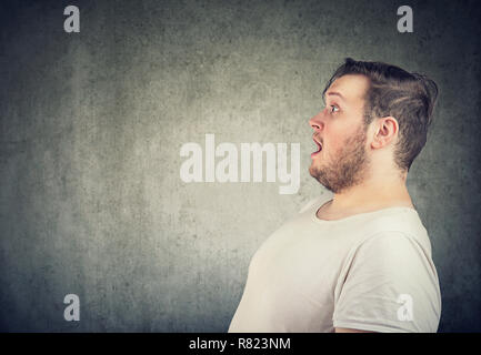 Vue de côté de l'homme grassouillet en t-shirt blanc à l'écart dans la stupéfaction avec bouche ouverte sur fond gris Banque D'Images