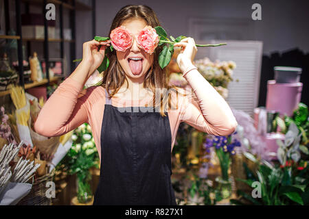 Drôle et positive young female florist couvrir les yeux avec des roses et de montrer la langue. Elle a l'amusement. Plantes et jardinières en pot derrière. Banque D'Images