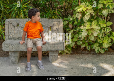 Un petit garçon concerné se trouve sur un banc de parc de ciment à la route. Il est habillé d'orange, t-shirt, short, TAN et gris shoes sit​ en attente pour maman Banque D'Images